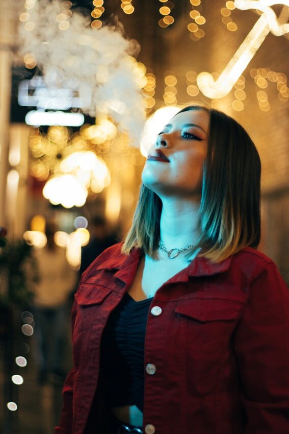 Stylish woman vaping in a brightly lit urban street, exhaling smoke surrounded by golden bokeh lights.