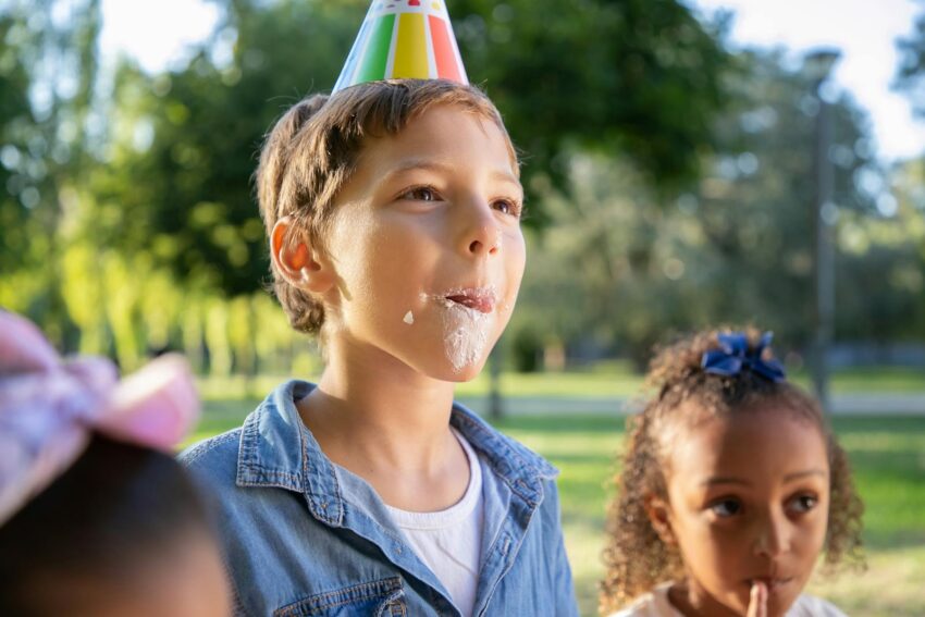 Children enjoying an outdoor birthday party with colorful hats and whipped cream treats.