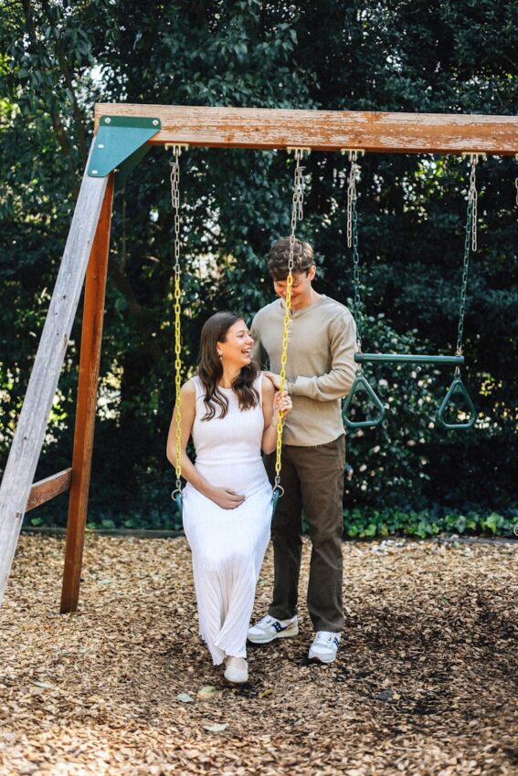 Pregnant couple poses together on a swing set.
