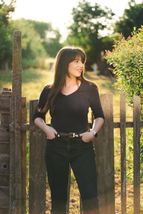 A woman wearing black poses by a rustic fence in a sunlit countryside scene.