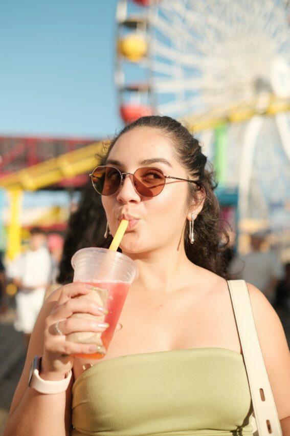 A woman in sunglasses drinking from a plastic cup at an amusement park