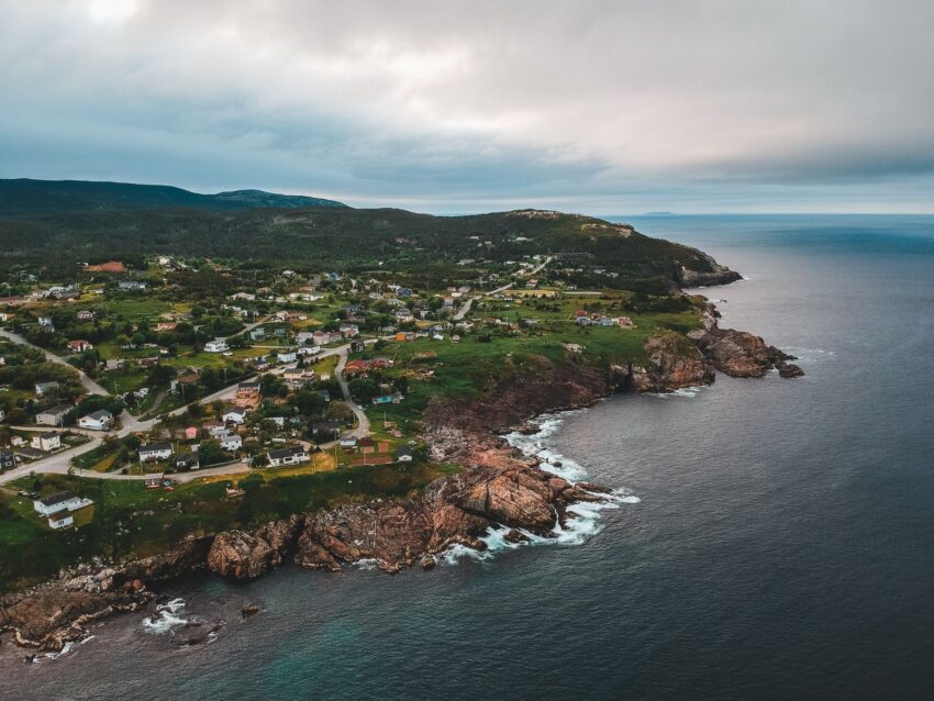 Drone view of rippled sea near lawns with dwelling buildings and mounts under shiny sky in evening