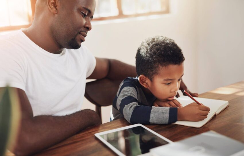 A father watches his son write in a notebook at a wooden table, with a tablet nearby.