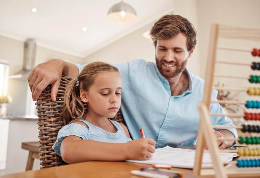 A father sits beside his daughter as she writes at a home table, with an abacus nearby.