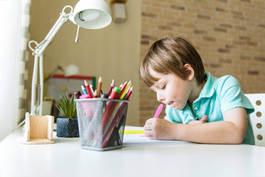 Boy drawing at a white desk beside a mesh cup of colored pencils and a desk lamp.